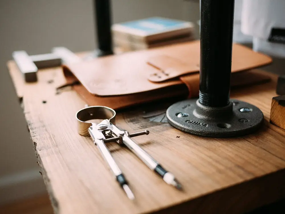 A pair of drawing dividers resting on a wooden bench top beside a leather folder