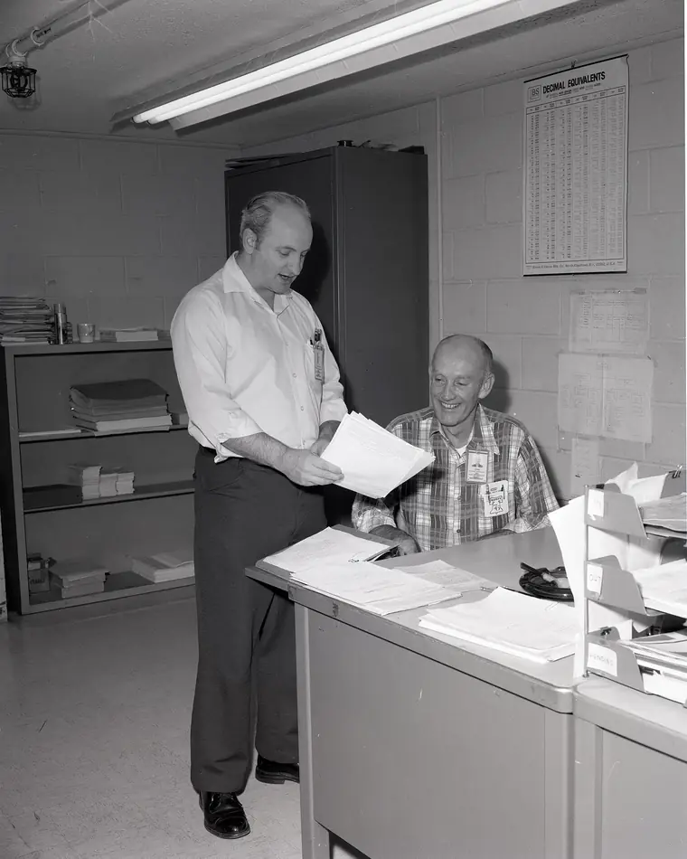A black-and-white photograph of a stockroom counter where two people check a paper list together