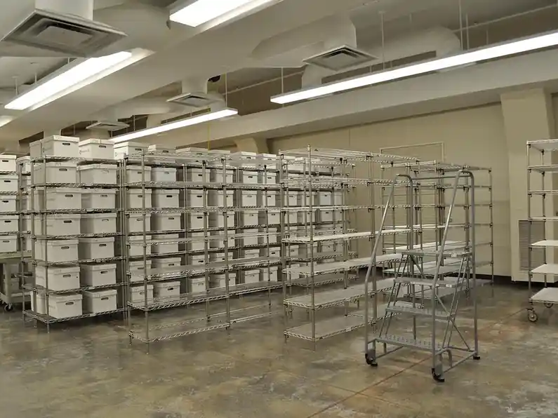 A long room of wire shelving units holding white archive boxes, with a rolling step ladder in the aisle